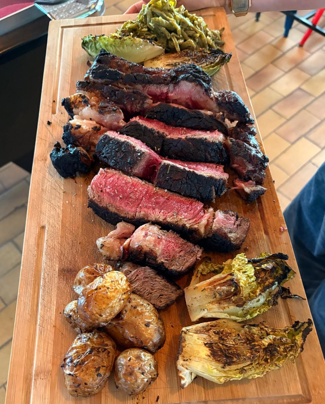 Côte de bœuf tranchée sur planche, croûte noire et chair rosée, avec pommes de terre grillées et sucrines à la braise